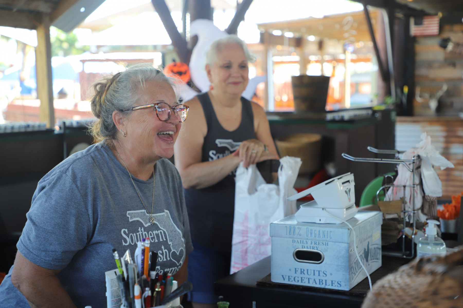 Two women stand behind a cashier's counter and are smiling and chatting with customers.